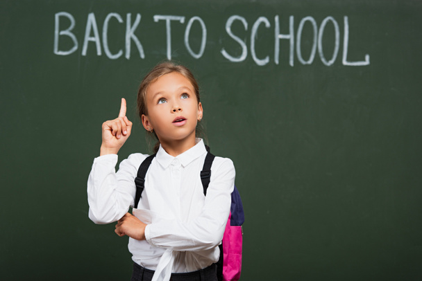 thoughtful schoolgirl showing idea gesture while looking up near chalkboard with back to school lettering - Фото, зображення
