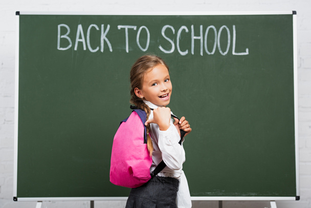 cheerful schoolgirl pointing with thumb at backpack near chalkboard with back to school lettering - Photo, Image