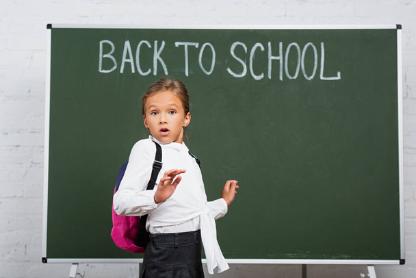 σοκαρισμένη μαθήτρια με σακίδιο κοιτάζοντας κάμερα κοντά στο chalkboard με πίσω στο σχολείο επιγραφή - Φωτογραφία, εικόνα