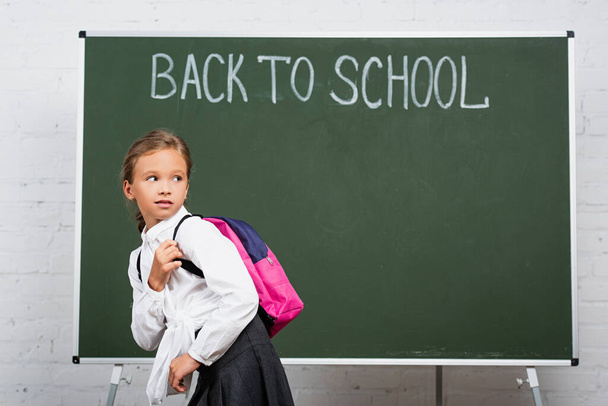 skeptical schoolgirl with backpack looking back near chalkboard with back to school inscription - Photo, Image