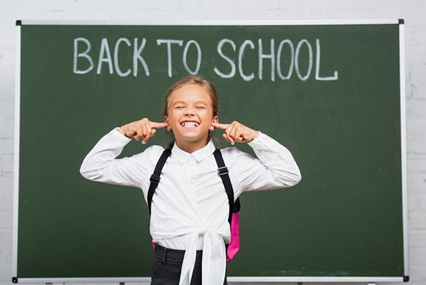 displeased schoolgirl plugging ears with fingers near chalkboard with back to school inscription - Photo, Image