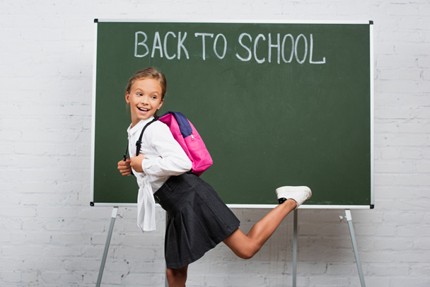 χαρούμενη μαθήτρια με σακίδιο στέκεται στο ένα πόδι κοντά στο chalkboard με πίσω στο σχολείο επιγραφή - Φωτογραφία, εικόνα