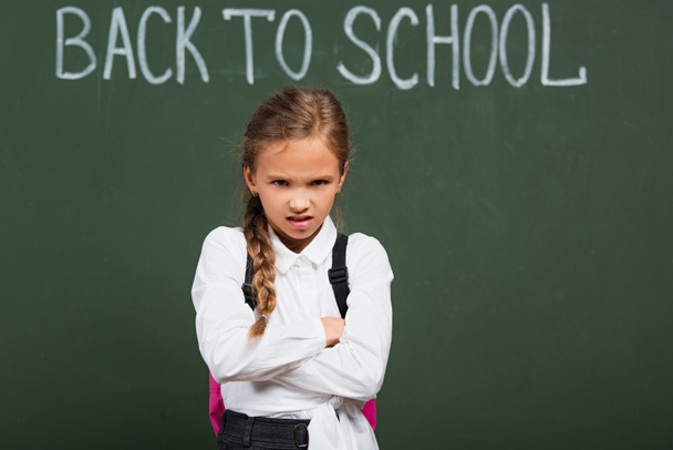 rozzlobený školačka stojící se zkříženýma rukama v blízkosti tabuli s zpět do školy písmo  - Fotografie, Obrázek
