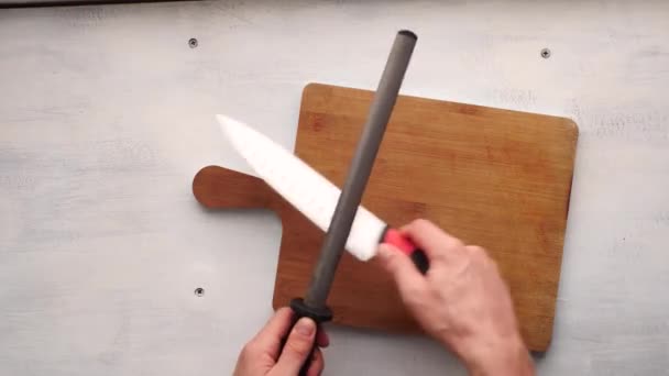 Білий чоловік наточує кухонний ніж з почесною сталлю над кліринговою дошкою в білому і рустиковому столику - Кадри, відео
