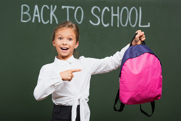 selective focus of smiling schoolgirl pointing at pink backpack near back to school lettering on chalkboard - Photo, Image