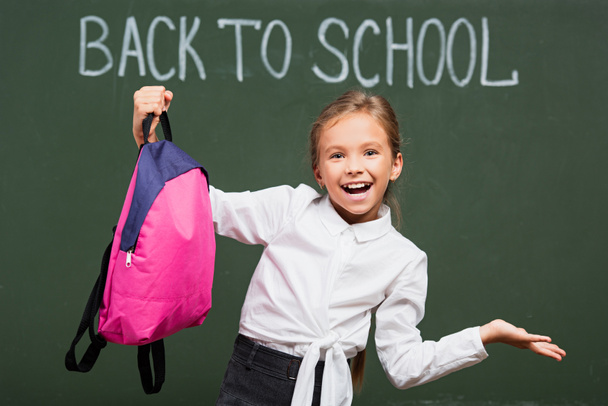colegiala alegre con el brazo abierto que sostiene la mochila cerca de pizarra con inscripción de vuelta a la escuela
 - Foto, Imagen