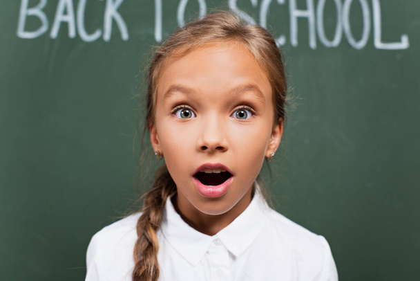 selective focus of shocked schoolgirl with open mouth looking at camera near chalkboard with back to school lettering - Photo, Image