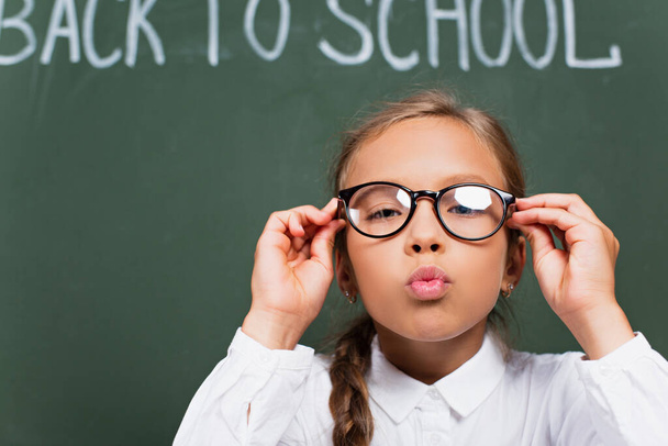 selective focus of cute schoolgirl touching eyeglasses and blowing air kiss near chalkboard with back to school lettering - Foto, Imagem