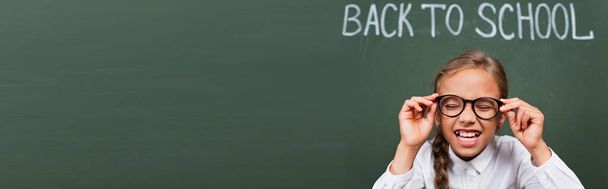 horizontal image of excited schoolgirl laughing with closed eyes and touching eyeglasses near chalkboard with back to school text - Photo, Image