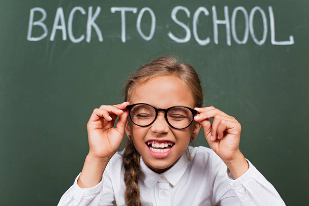 selective focus of excited schoolgirl laughing with closed eyes and touching eyeglasses near chalkboard with back to school inscription - Photo, Image