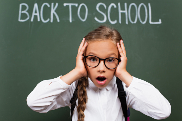 selective focus of shocked schoolgirl in eyeglasses touching head near back to school lettering on chalkboard - Foto, Imagem