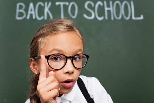 selective focus of adorable schoolgirl in eyeglasses showing idea gesture near chalkboard with back to school text - Photo, Image