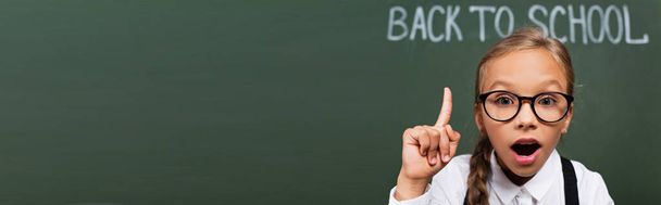 horizontaal beeld van verrast schoolmeisje met idee gebaar in de buurt van schoolbord met terug naar school belettering - Foto, afbeelding