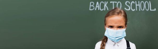 horizontal image of displeased schoolgirl in protective mask looking at camera near back to school inscription on chalkboard - 写真・画像