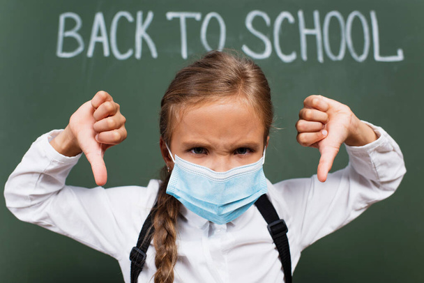 selective focus of displeased schoolgirl in medical mask showing thumbs down near chalkboard with back to school inscription - Foto, imagen