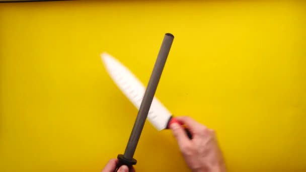 La main de l'homme blanc affûtant un couteau de cuisine avec un acier à aiguiser sur un fond jaune - Séquence, vidéo