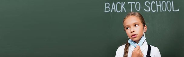 horizontal concept of tired schoolgirl taking off protective mask near chalkboard with back to school lettering - Photo, Image