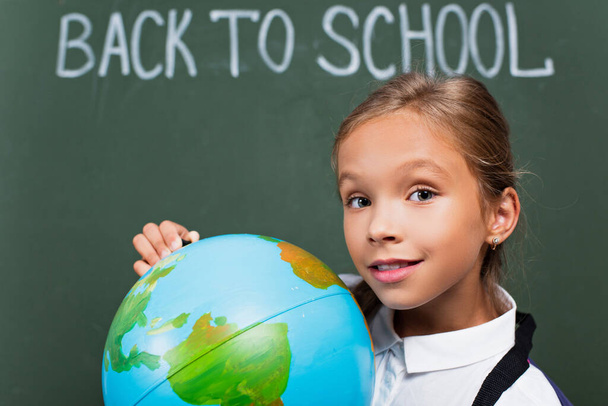 selective focus of smiling schoolgirl holding globe and looking at camera near back to school inscription on chalkboard - Photo, Image