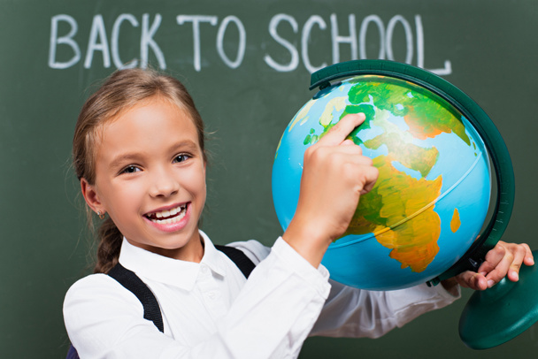 selective focus of happy schoolgirl pointing with finger at globe near back to school inscription on chalkboard - Фото, изображение
