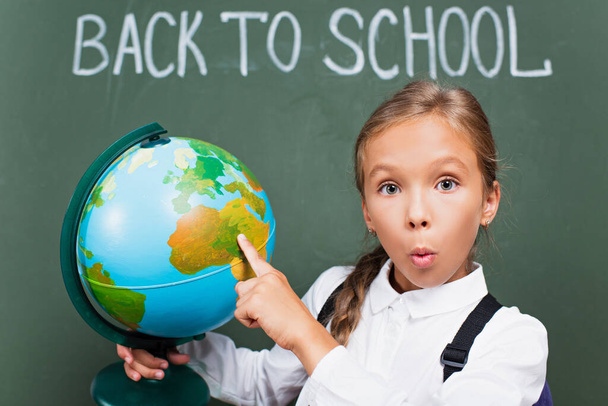 surpris écolière pointant avec doigt au globe près de retour à l'école lettrage sur tableau noir - Photo, image