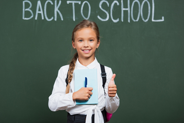 cheerful schoolgirl with book and pen showing thumb up near chalkboard with back to school lettering - Photo, Image
