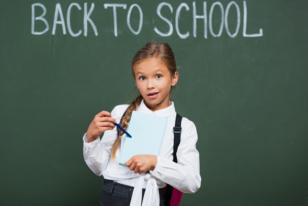 écolière surprise pointant avec stylo au livre près du tableau noir avec inscription de retour à l'école - Photo, image