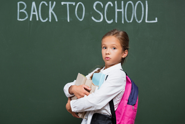 discouraged schoolgirl with backpack holding books near chalkboard with back to school inscription - Foto, Imagem
