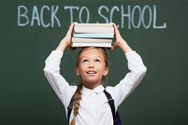 selective focus of smiling schoolgirl holding stack of books above head near chalkboard with back to school text - Photo, Image