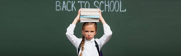récolte panoramique d'écolière mécontente tenant pile de livres au-dessus de la tête près du tableau noir avec lettrage de retour à l'école - Photo, image