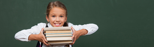 horizontal image of happy schoolgirl showing stack of books near green chalkboard - 写真・画像