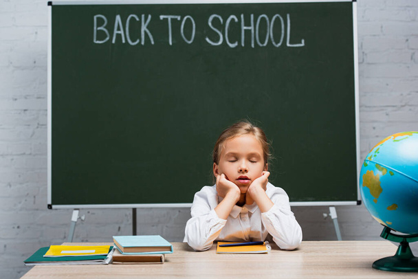 tired schoolgirl sitting with closed eyes at desk near globe and chalkboard with back to school inscription - Foto, Imagem