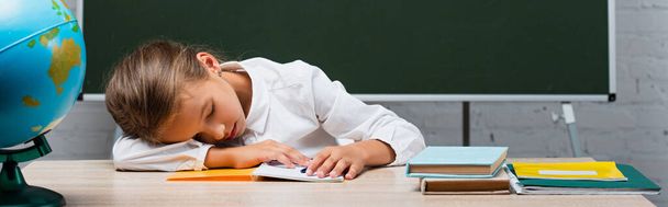 imagem horizontal de estudante cansada dormindo na mesa perto do globo e quadro-negro com as costas para a inscrição da escola
 - Foto, Imagem