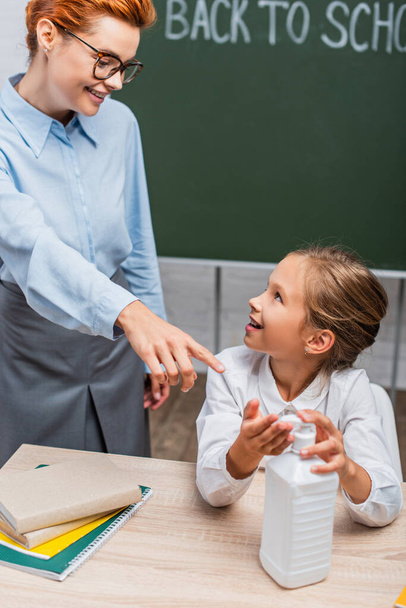 selective focus of smiling teacher pointing with finger near schoolgirl applying hand antiseptic  - Фото, изображение