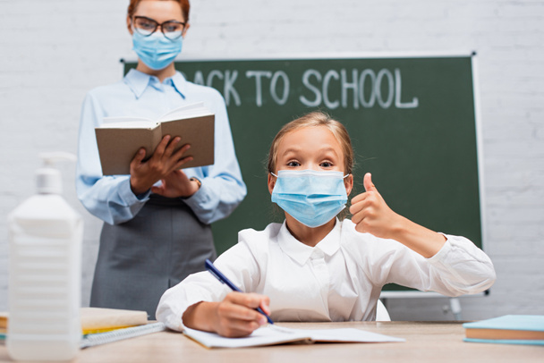 selective focus of schoolgirl in protective mask showing thumb up, and teacher standing near chalkboard with back to school lettering - Foto, Imagem