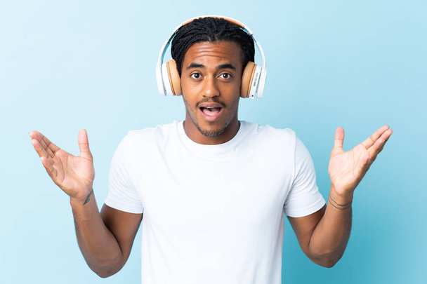 Young African American man with braids isolated on blue background surprised and listening music - Photo, Image