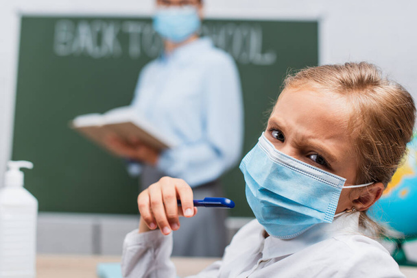 selective focus of frowning schoolgirl in medical mask holding pen and looking at camera, while teacher standing near chalkboard  - Фото, изображение