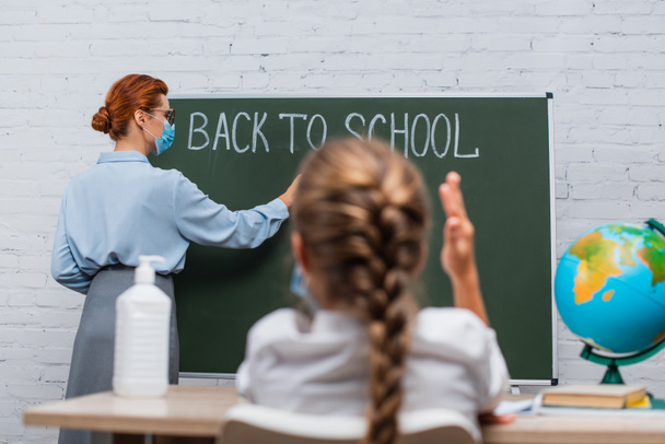 visszapillantás iskolás lány emelt kézzel, és tanár orvosi maszk írás táblán vissza az iskolai betű - Fotó, kép