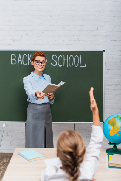 back view of schoolgirl with raised hand and smiling teacher near chalkboard with back to school lettering - Foto, afbeelding