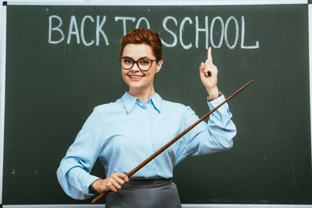 smiling teacher holding pointer and pointing with finger at back to school inscription on chalkboard - Foto, immagini