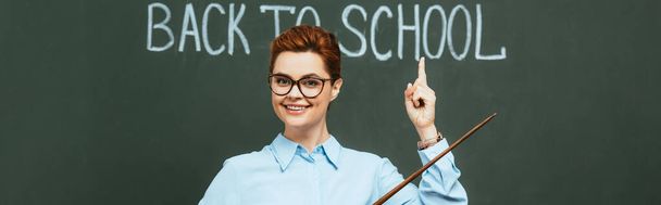Panoramaaufnahme eines glücklichen Lehrers, der Zeiger hält und mit dem Finger auf den Schulschriftzug auf der Tafel zeigt - Foto, Bild