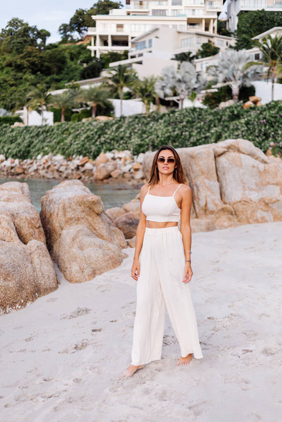 Romantic portrait of young calm happy caucasian fit slim woman in crop cami top and pants set sits alone on rocky tropical beach at sunset. Luxury villas on background. - Fotoğraf, Görsel