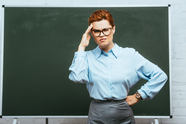 exhausted teacher with headache touching forehead while standing with hand on hip near chalkboard - Foto, Imagen