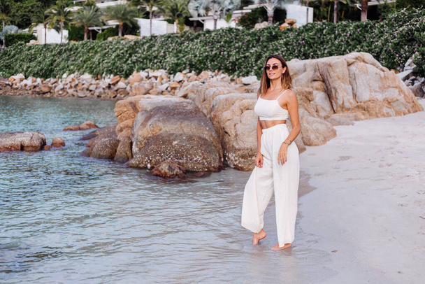 Romantic portrait of young calm happy caucasian fit slim woman in crop cami top and pants set sits alone on rocky tropical beach at sunset. Luxury villas on background. - Valokuva, kuva