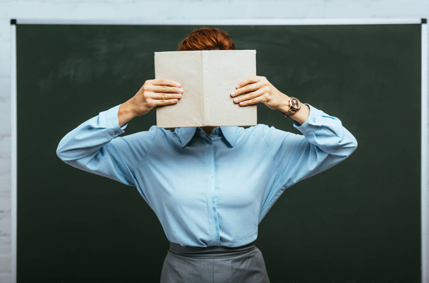 teacher in blue blouse obscuring face with book while standing near chalkboard  - Foto, afbeelding