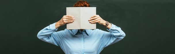 panoramic crop of teacher obscuring face with open book while standing near chalkboard  - Fotoğraf, Görsel