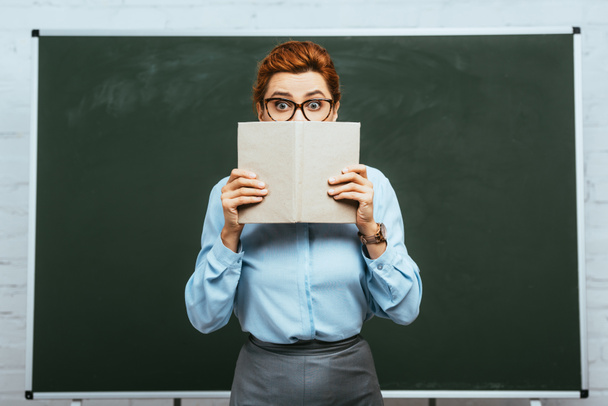 scared teacher in eyeglasses covering face with open book while standing near chalkboard  - Photo, Image