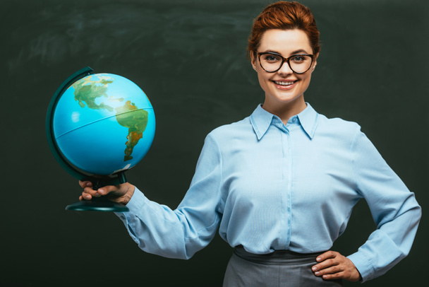 Glückliche junge Lehrerin steht neben Kreidetafel, die Hand auf der Hüfte und hält Globus - Foto, Bild