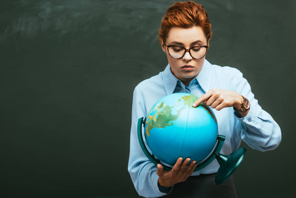 attractive young teacher pointing with finger at globe while standing near chalkboard  - Foto, Imagen