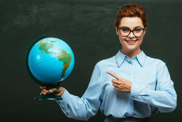 Lächelnder Lehrer zeigt mit dem Finger auf Globus, während er neben Tafel steht  - Foto, Bild