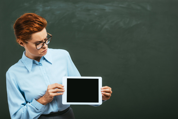 вдумчивый учитель смотрит на цифровой планшет с чистым экраном рядом с доской - Фото, изображение
